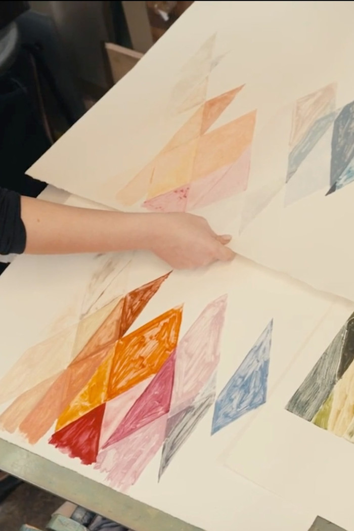 A person holds large sheets of paper displaying hand-painted geometric patterns in various colours, including orange, pink, blue and yellow.