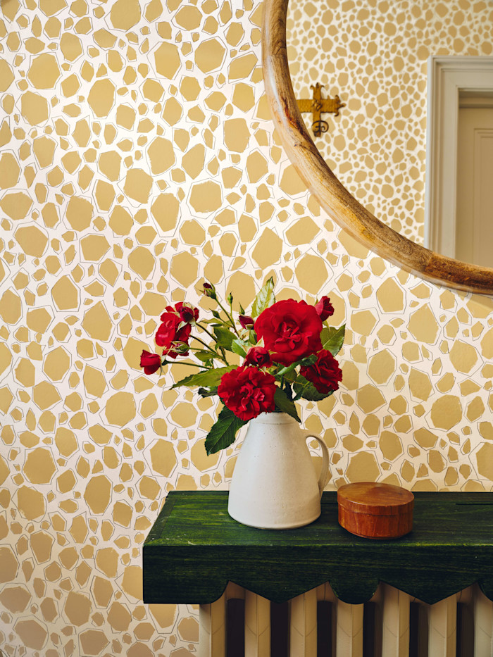 A white ceramic pitcher with red flowers sits on a dark green shelf against a patterned beige and white wall, with a round mirror above.