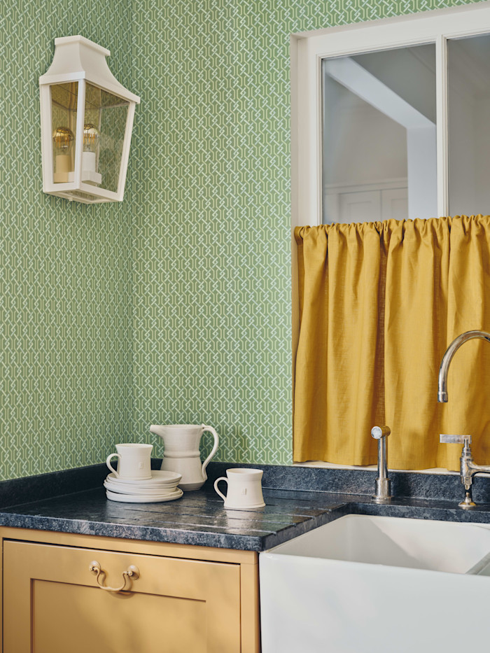 A kitchen corner with green geometric-patterned wallpaper, yellow cabinetry, a white farmhouse sink, and a mustard curtain over the window. Stacked white dishes, mugs and a pitcher sit on a dark countertop.