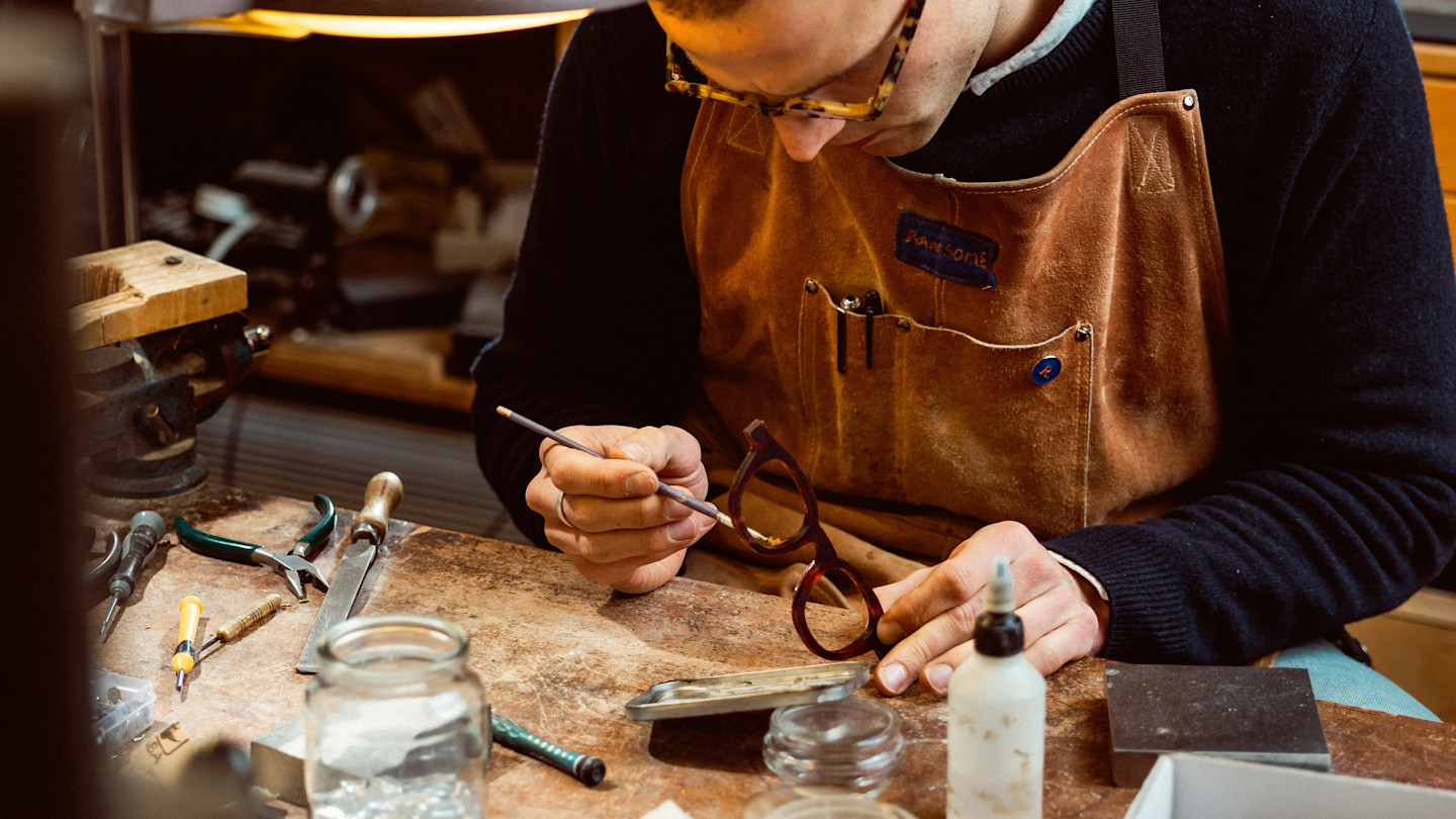A young man in a suede apron leans over a workbench, holding a pair of glasses.