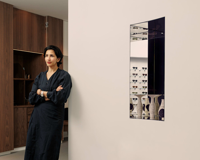 Ahlem Manai-Platt stands with arms crossed in a minimalist room, beside a wall with a mirror reflecting shelves of sunglasses.