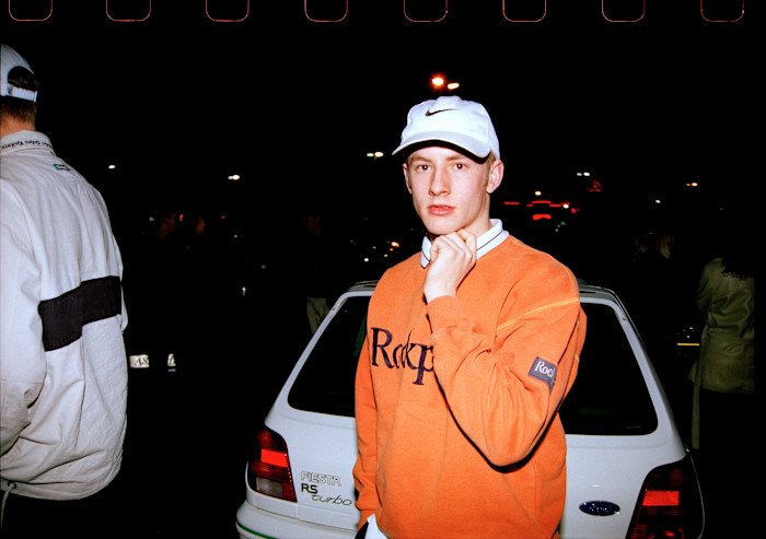 A young man in a white baseball cap and orange sweatshirt poses beside a white hatchback car at night.