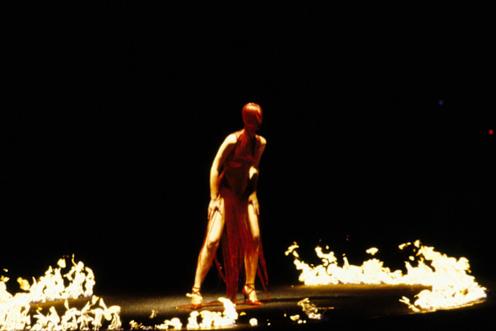 A model in a red dress and head covering stands inside a ring of fire on a dark runway at Alexander McQueen’s AW 1998 show.