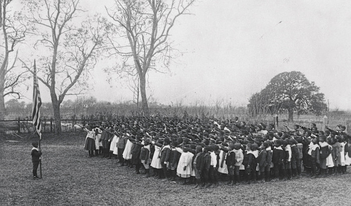 A large group of schoolchildren stand outdoors, facing a child holding an American flag, with arms raised in salute.