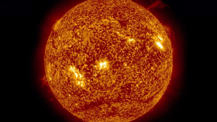 A close-up view of the sun, showing its textured, fiery surface with bright spots and a glowing red aura against a black background.