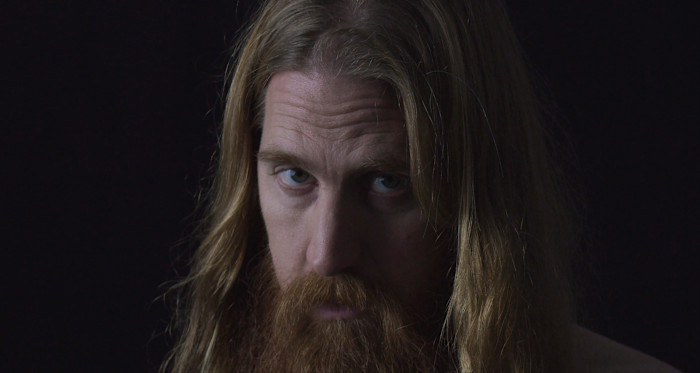A person with long hair and a full beard looks directly at the camera against a dark background.