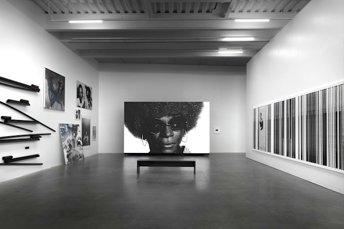 Installation view of "Arthur Jafa: I Am Tony" at the New Museum, showing a large black-and-white portrait of a person with an afro and sunglasses, surrounded by various artworks on the walls.