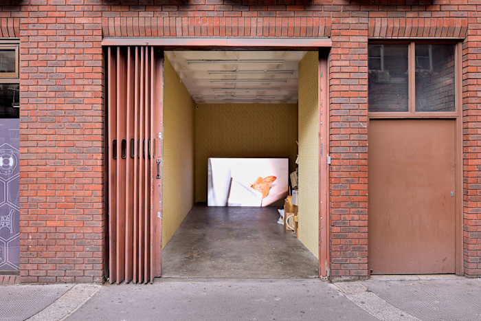 A small brick storefront with an open folding door reveals Minne Kersten's artwork "room temperature," featuring a large illuminated photograph of a moth inside.