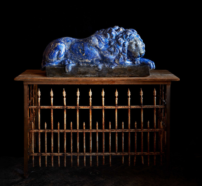 Blue reclining lion sculpture displayed on top of a rustic iron console with spear-like vertical bars.