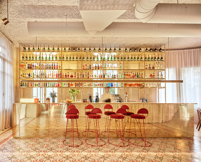 A bar with a mirrored counter, red barstools, and shelves filled with colourful bottles and glassware. A person stands behind the counter.
