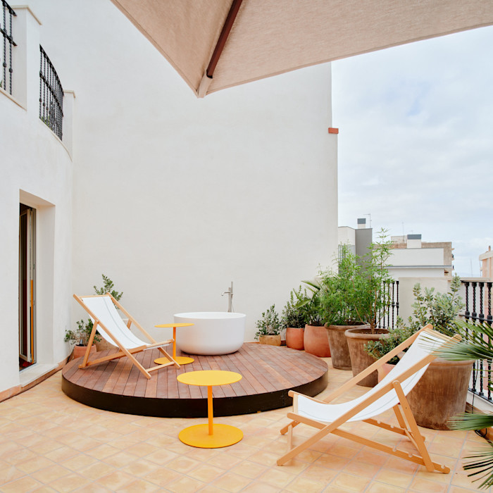 Two white deck chairs with wooden frames and two small round yellow tables are arranged on a tiled terrace. A round wooden platform holds a white outdoor tub surrounded by potted plants. An umbrella provides shade from above.