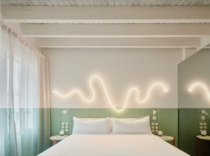 A minimalist bedroom with a white bed, green accent wall, and wavy LED light fixture above the headboard.