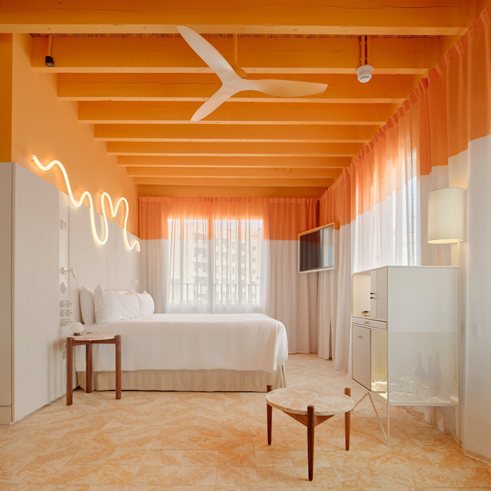 A hotel room with a white bed, peach and white curtains, a ceiling fan, and modern minimalist furniture under a wooden beam ceiling.