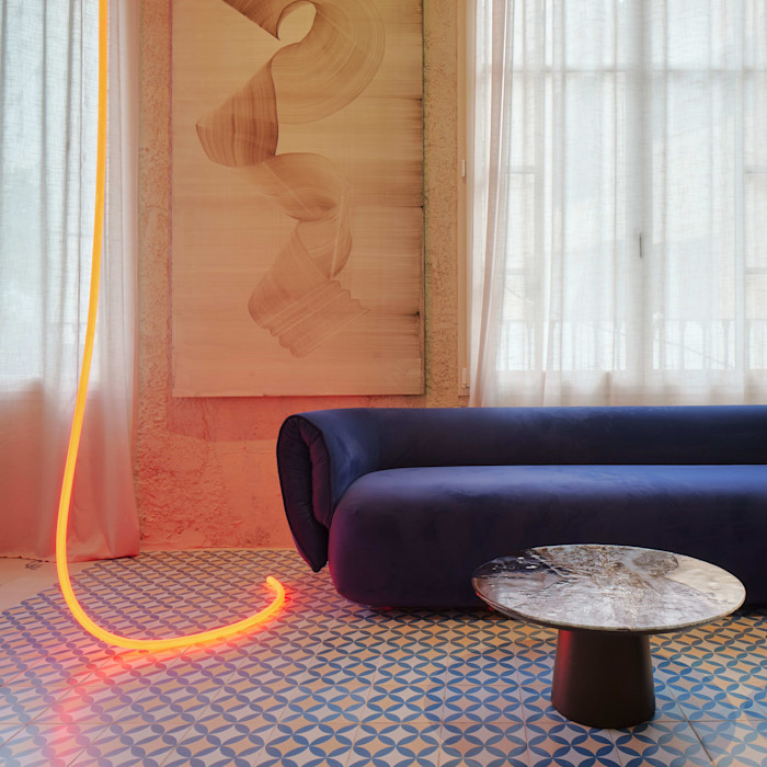 A hotel room with a dark blue minimalist sofa, toadstool-style coffee table, swirling figurative picture on the wall and a long orange LED rope-like light dangling on the floor.