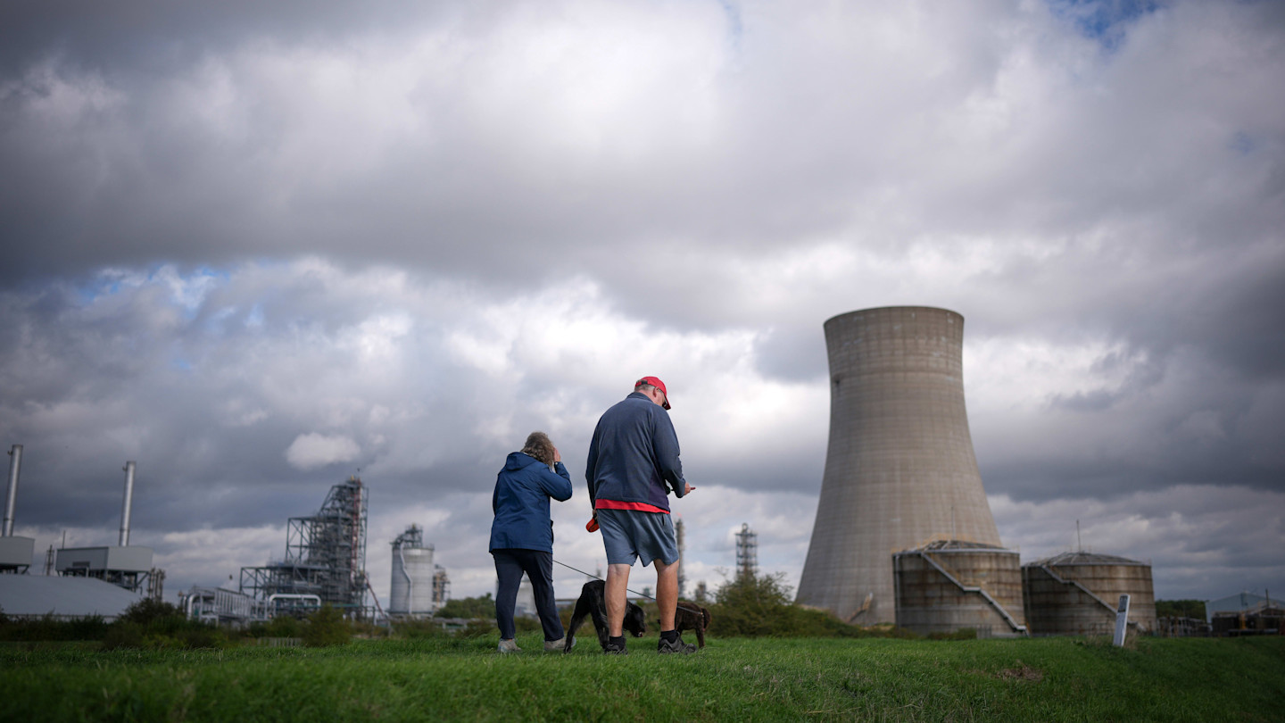 Two people walk dogs on a grassy area near large industrial structures and a cooling tower under a cloudy sky.