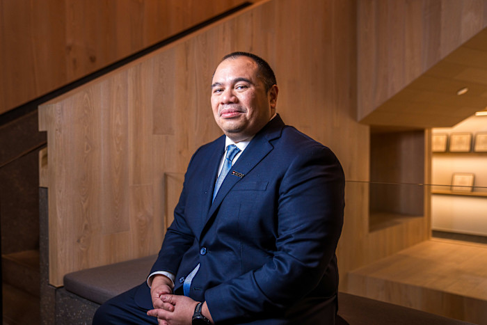 Pandu Sjahrir seated indoors in a business suit, looking at the camera, with wooden walls and stairs in the background.