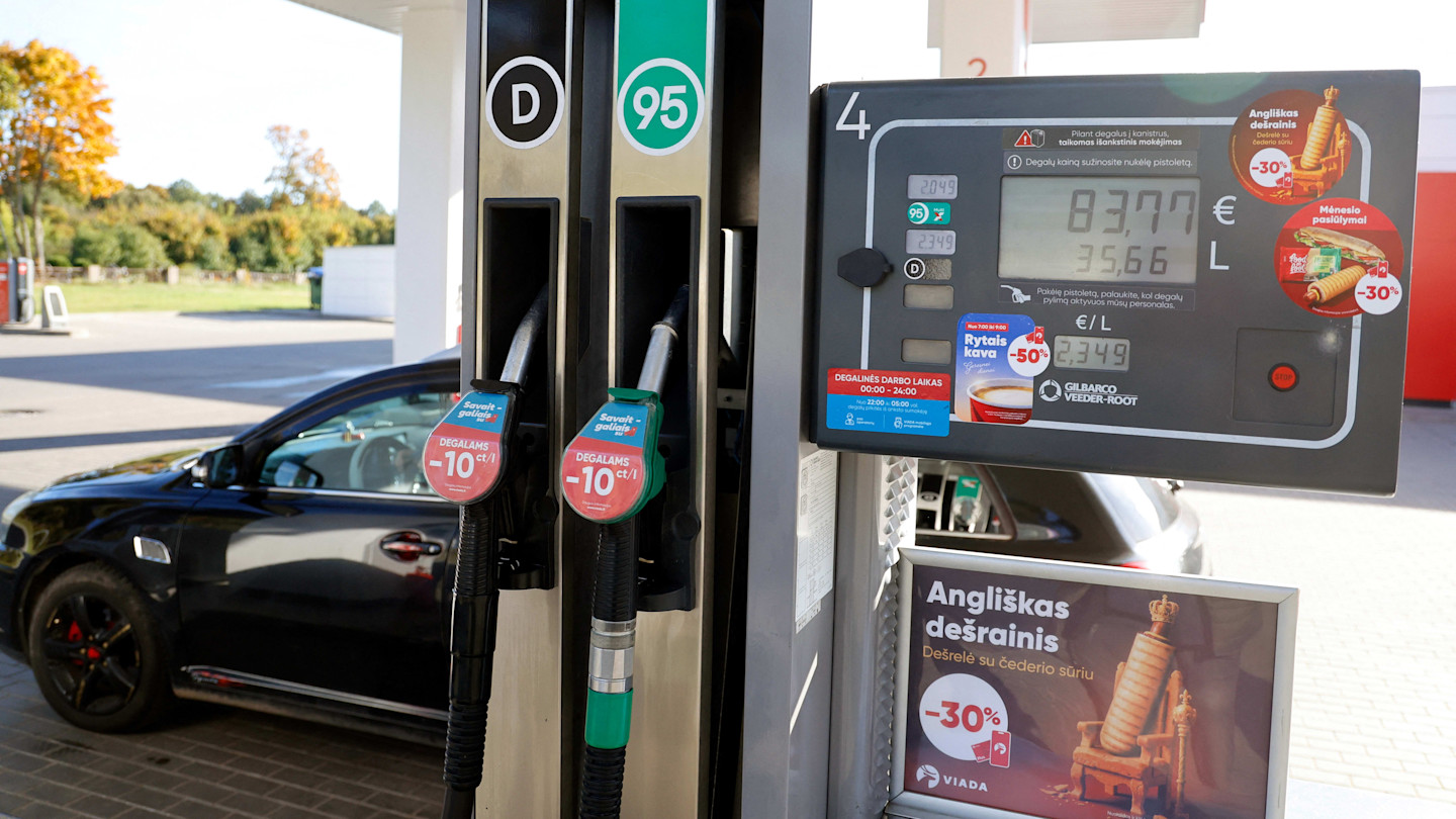 A fuel pump at a petrol station in Varena, Lithuania, displays fuel prices and promotional stickers, with a black car parked beside it.