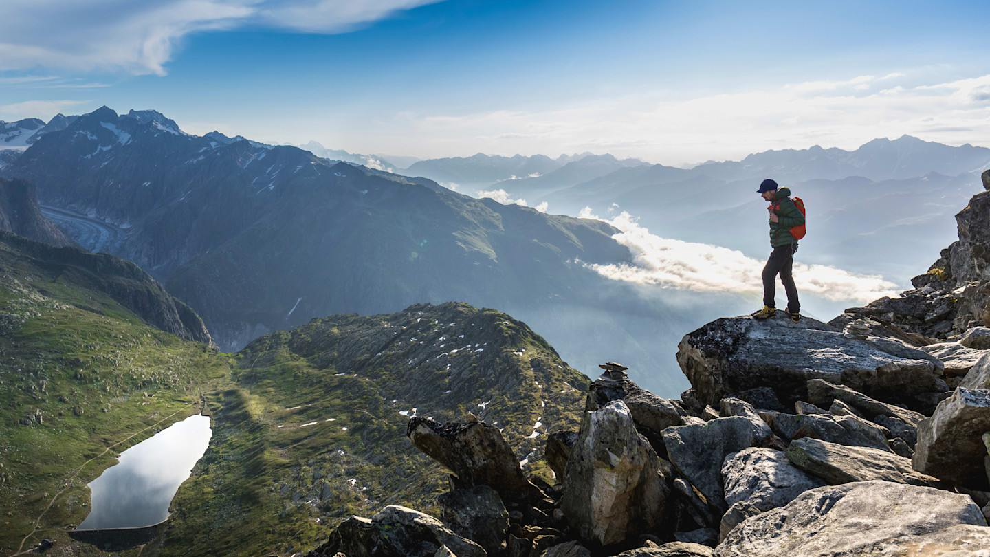 A hiker stands on a rocky outcrop overlooking a panoramic mountain landscape with distant peaks and a lake below.