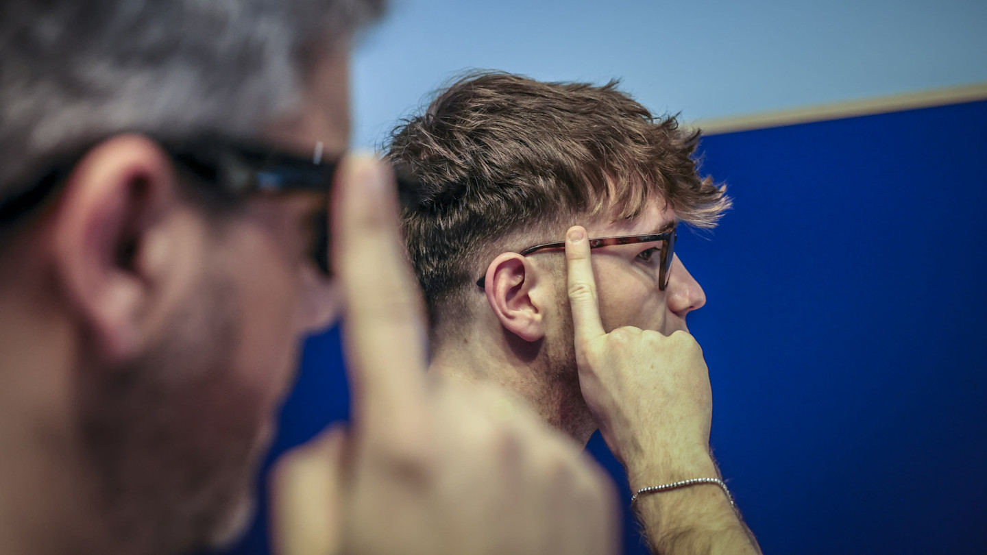 Two attendees try on Ray-Ban Meta AI glasses, each touching the side of the frames, with a blue background.