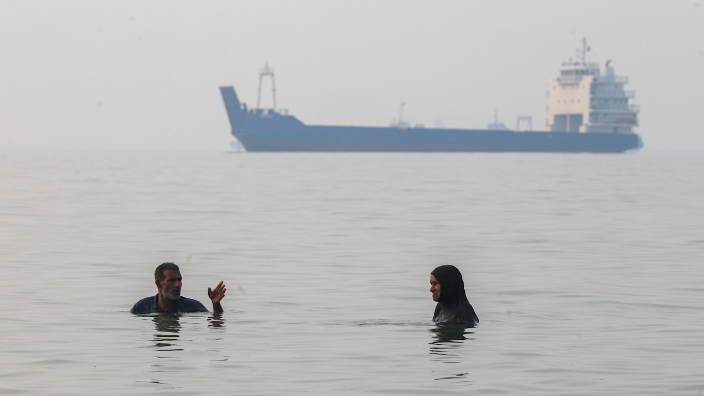 A man and a woman stand waist-deep in water, with a large commercial ship anchored in the hazy distance behind them.