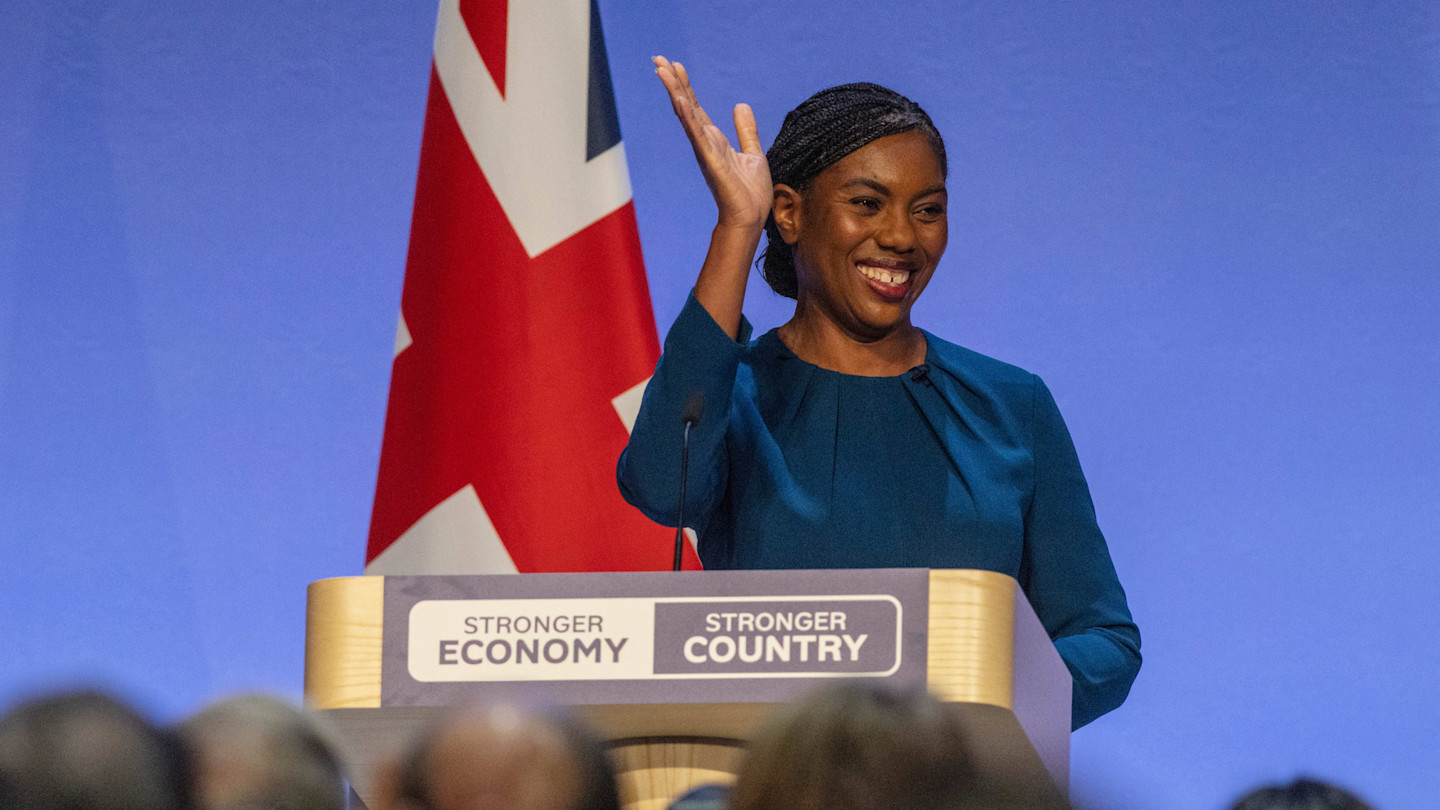 Kemi Badenoch stands at a podium, smiling and waving, with a Union Jack flag in the background.