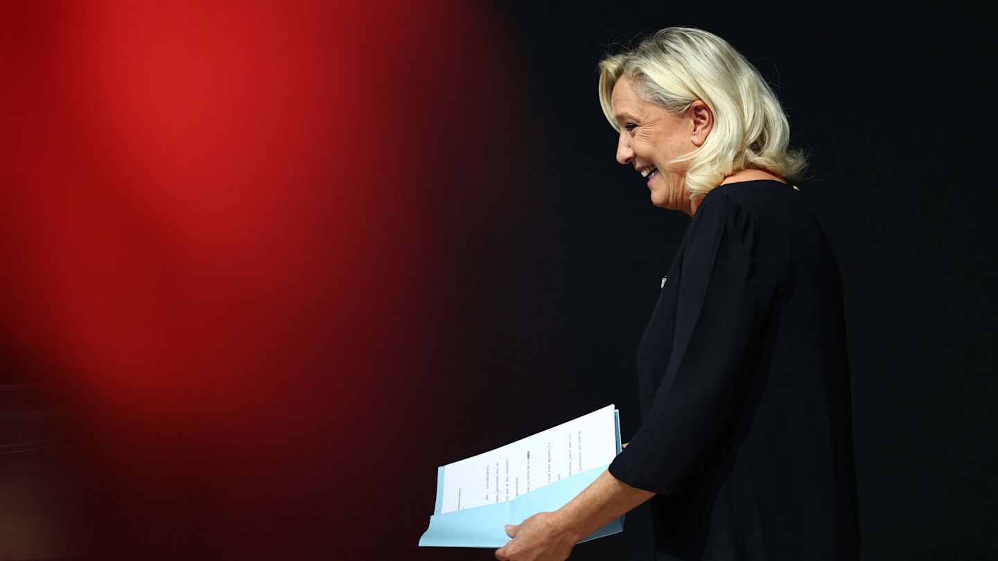 Marine Le Pen stands in profile holding documents, smiling, against a dark background with a red blur on the left.