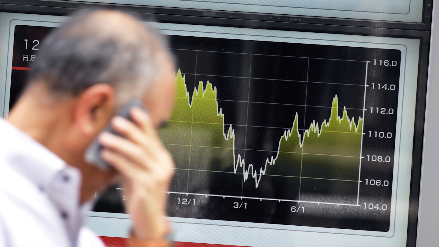 A person on a phone walks past an electronic board showing a fluctuating graph