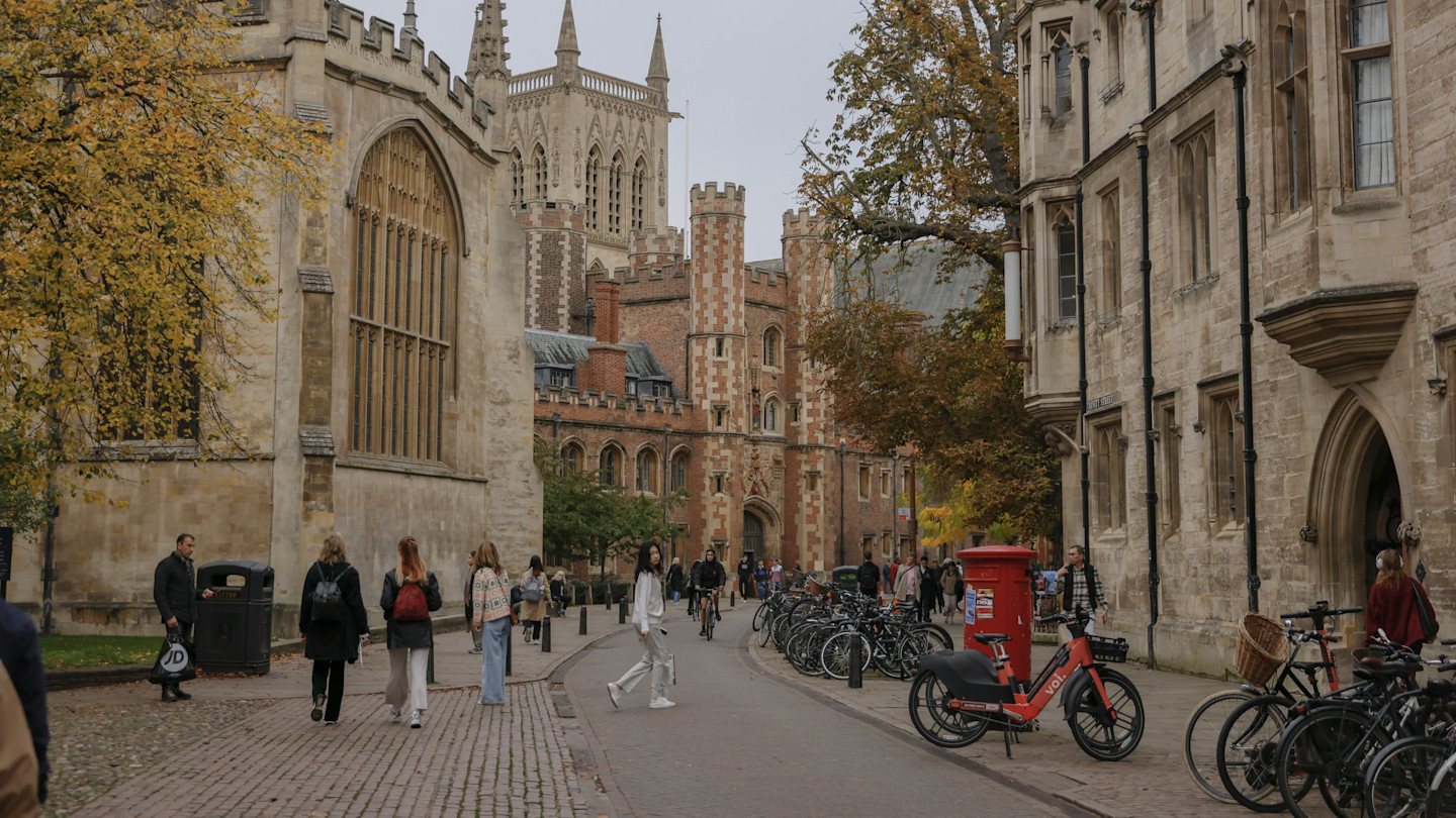 Pedestrians and cyclists move along a cobblestone street lined with historic college buildings and bicycles near St John’s College, Cambridge.