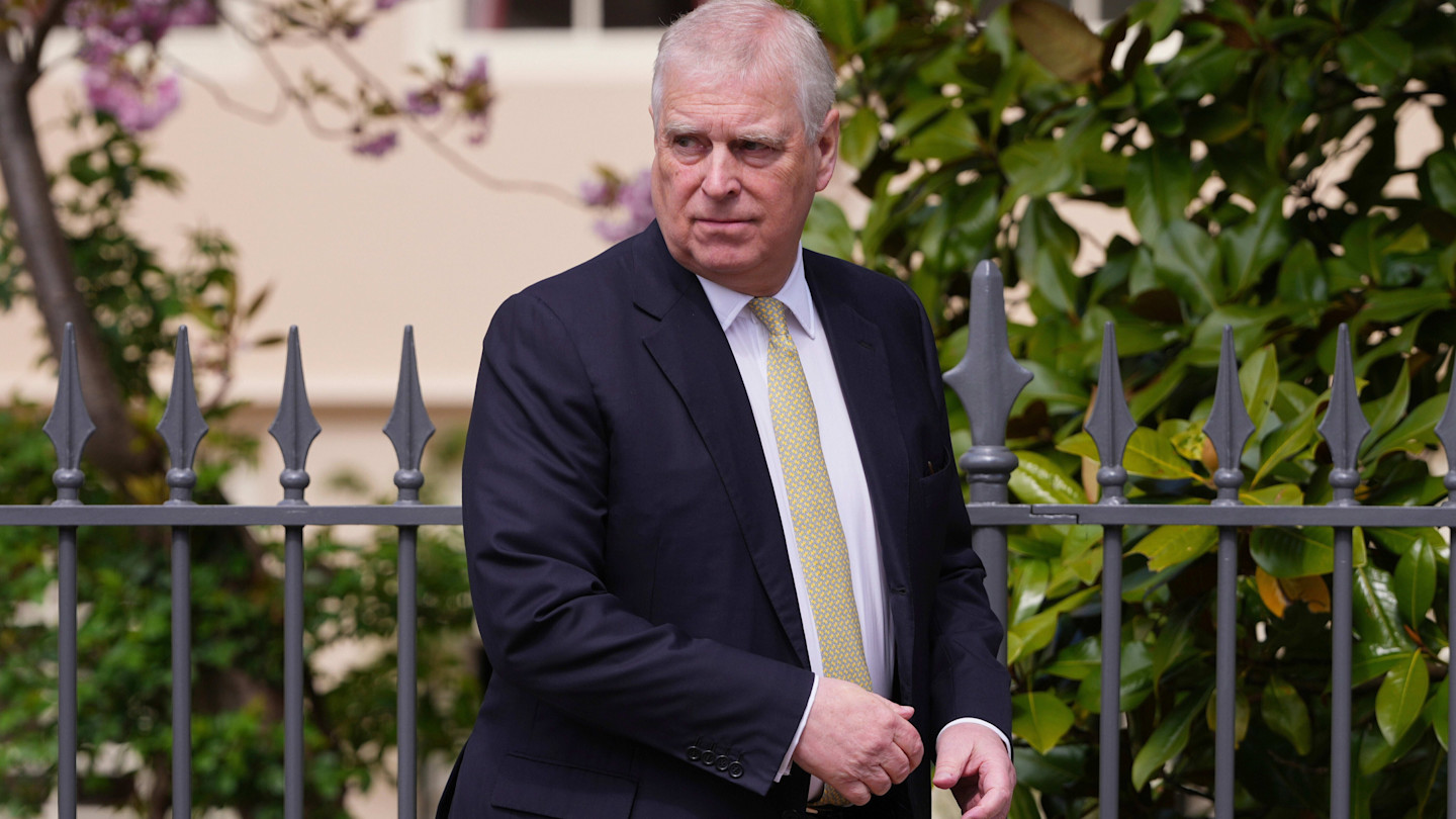 Andrew Mountbatten-Windsor, formerly known as Prince Andrew, stands outside near an iron fence, wearing a dark suit and yellow tie.