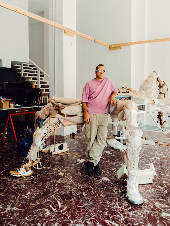 Theo Triantafyllidis stands in his Athens studio, leaning on his sculpture “Feral Metaverse (Spider),” surrounded by art materials.