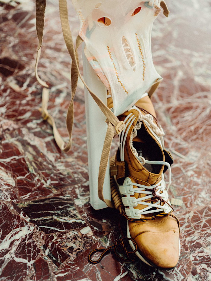 A close-up of part of Theo Triantafyllidis’s sculpture “Feral Metaverse (Spider),” showing a tan shoe with white plastic elements and a mask-like structure with stitched openings, on a marbled floor.