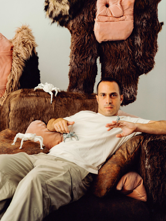 Theo Triantafyllidis reclines on a brown, fur-covered couch in his studio, with small sculptural creatures on his lap and beside him.