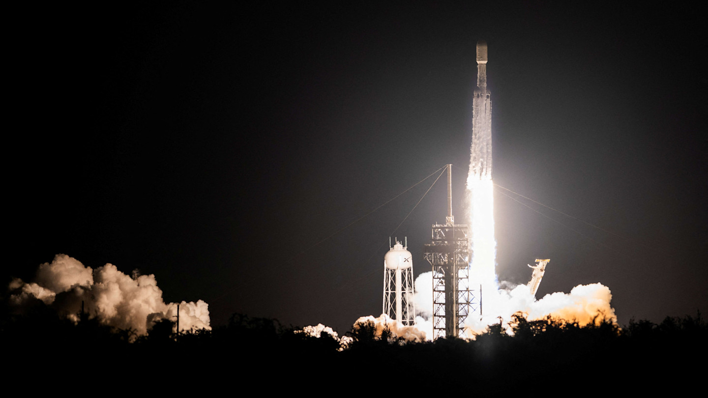 SpaceX Falcon Heavy rocket launches at night, with bright flames and smoke billowing from the pad during lift-off.