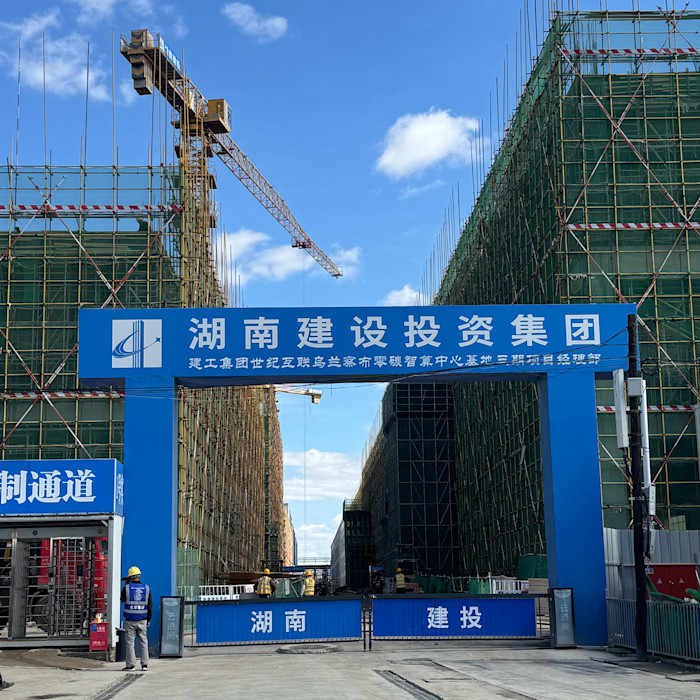 A large construction site with scaffolding on two buildings and a crane overhead. Workers and a blue sign with Chinese text are visible at the entrance.