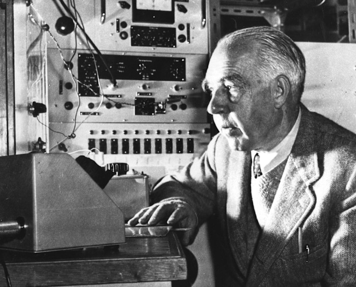 A black-and-white photograph of a man in late middle age, wearing a tweed jacket and tie, in front of a panel of dials and switches.