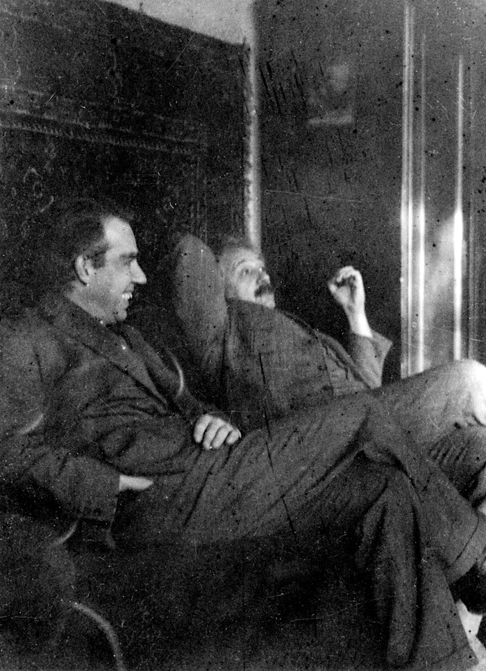 A blurred black-and-white photograph of Niels Bohr and Albert Einstein sit together, both appearing relaxed and engaged in conversation.