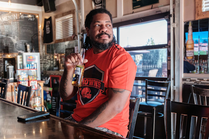 Robert Hines sits at a bar holding a bottle of beer, wearing a red Browns shirt and looking to the side.
