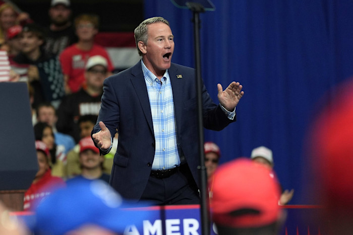 Senator Jon Husted gestures with open hands while speaking at a rally, with a crowd and American flag in the background.