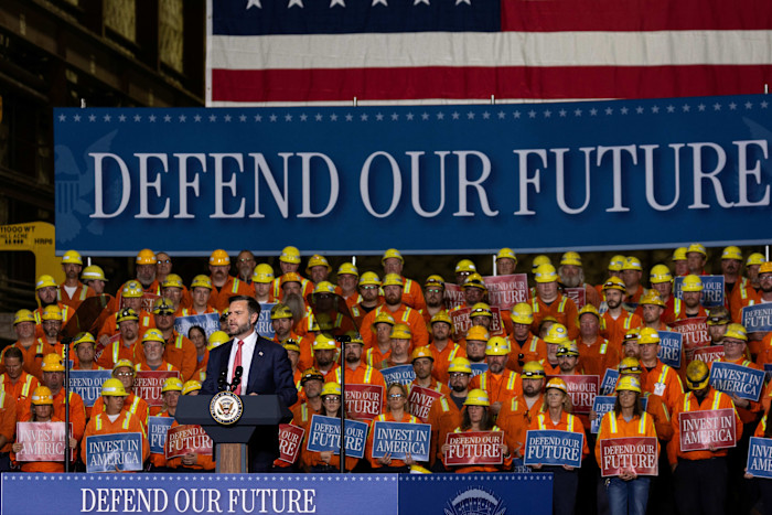 JD Vance speaks at a podium in front of steelworkers in orange uniforms and yellow helmets holding ‘Defend Our Future’ and ‘Invest in America’ signs