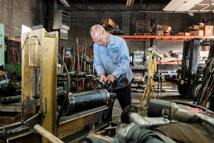 James Leicher uses a power tool to repair hydraulic equipment in his workshop, surrounded by machinery and tools.