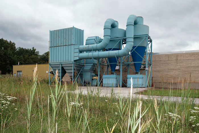 Exterior of the former Conn Selmer plant with overgrown grass and large blue industrial ventilation equipment.