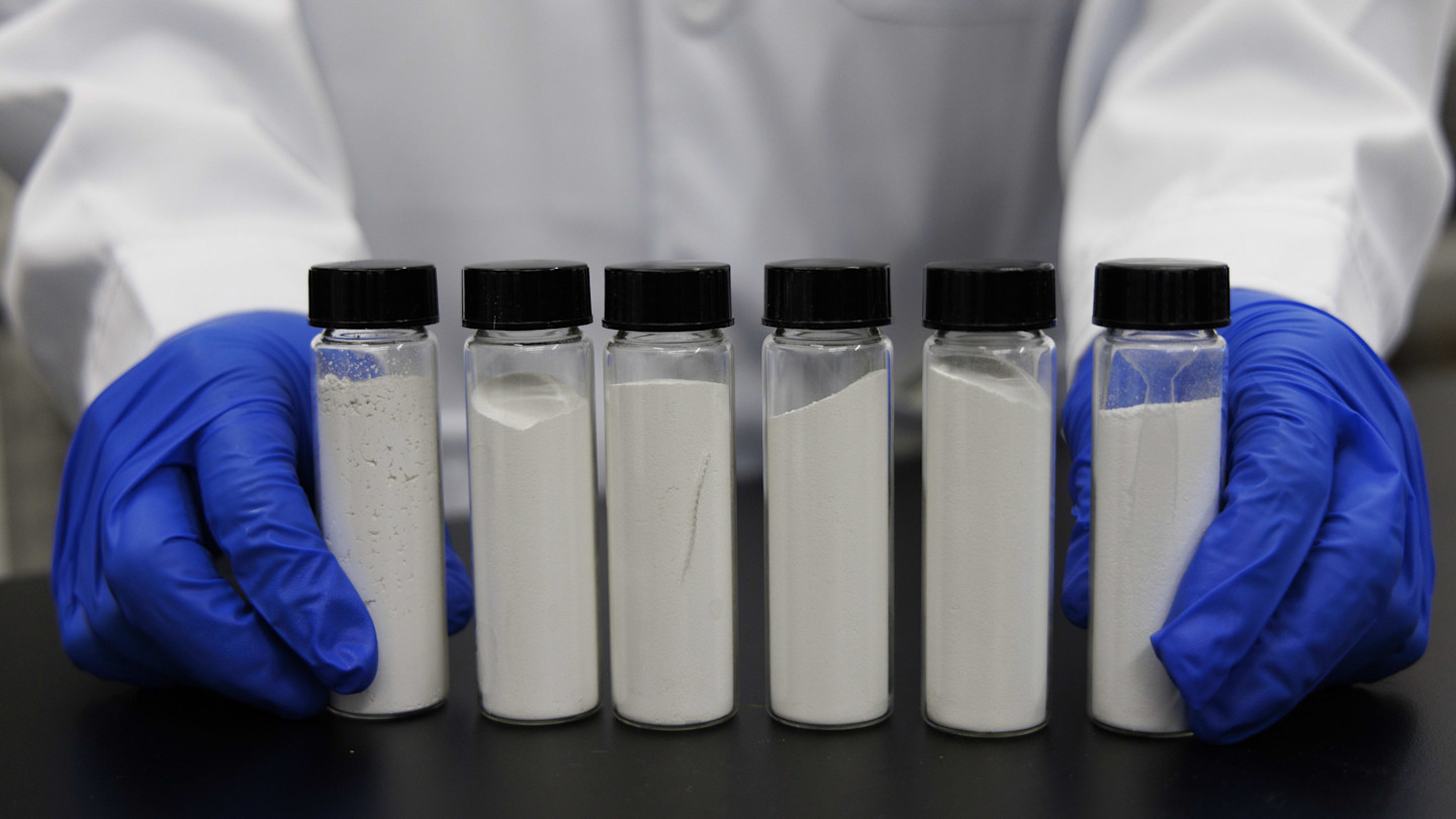 Six glass vials containing white titanium dioxide powder are displayed on a table, held by a researcher wearing blue gloves and a white lab coat.
