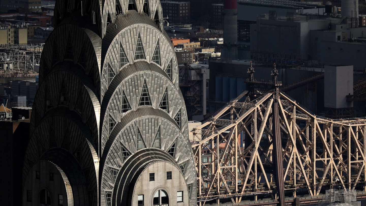 Close-up of the Chrysler Building’s Art Deco crown with the Queensboro Bridge and city buildings in the background.