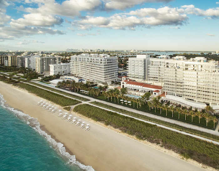 Aerial view of the Four Seasons Hotel at The Surf Club, Miami, with modern beachfront buildings, palm trees, and rows of white umbrellas on the sandy shore.