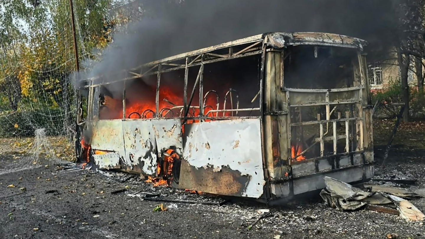 A burnt-out bus with flames and smoke rising from inside.