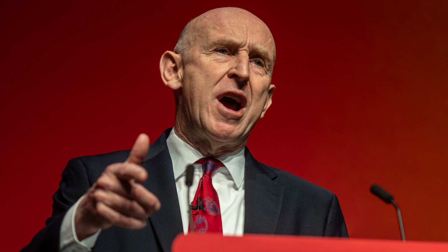 John Healey speaks at a podium, gesturing with his hand during his keynote speech against a red background.