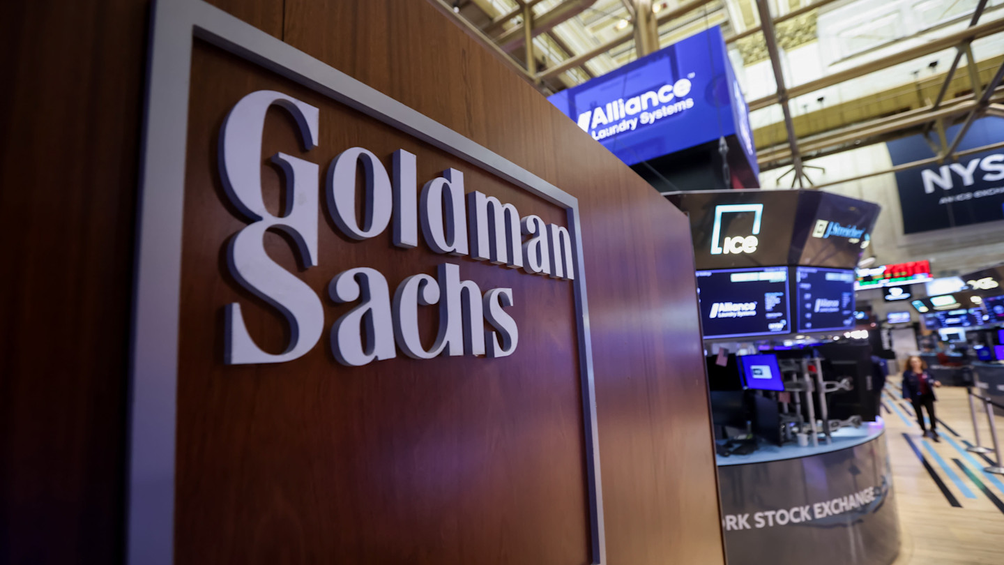 A Goldman Sachs sign on a wood panel at the New York Stock Exchange trading floor, with monitors and trading stations in the background.