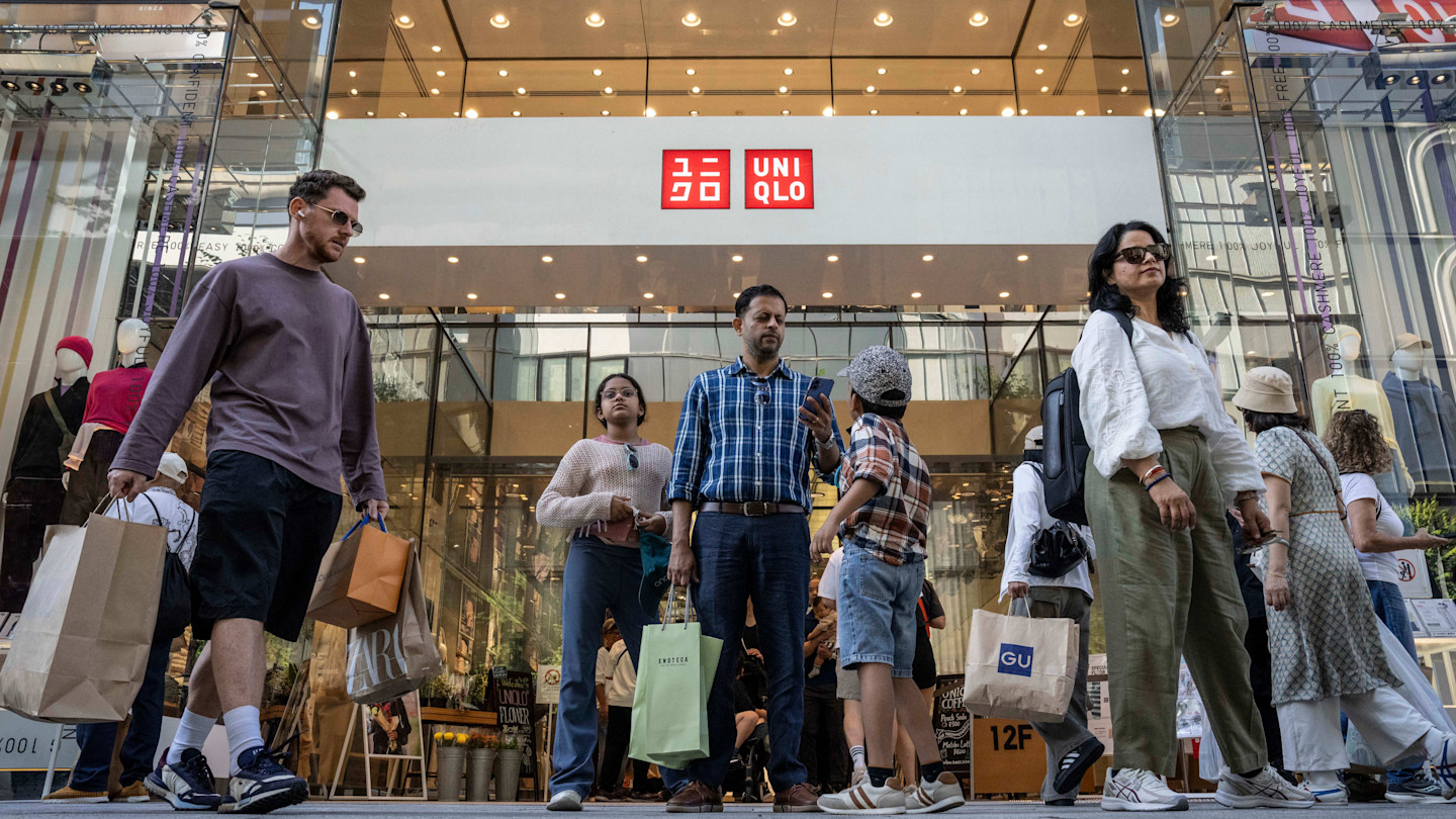 Shoppers carrying bags walk out of a Uniqlo store in Tokyo, with mannequins visible through the glass storefront.