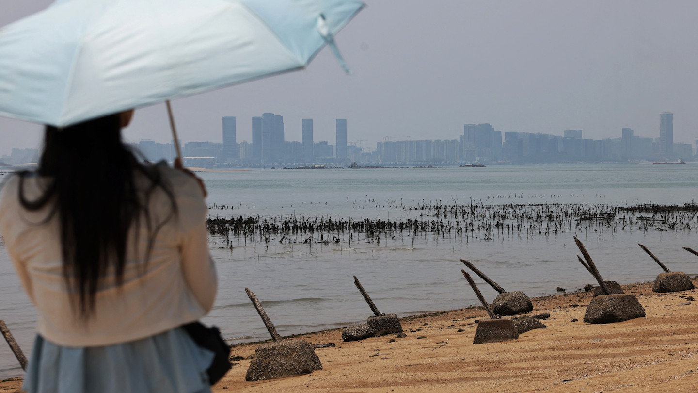 A person holding a light blue umbrella stands on a beach on Kinmen island, looking across the water toward the skyline of Xiamen, China. Concrete barriers are visible on the sand.