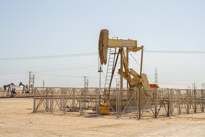 A pump jack extracts oil from a fenced well site in the Bahrain desert, with additional pump jacks visible in the background.